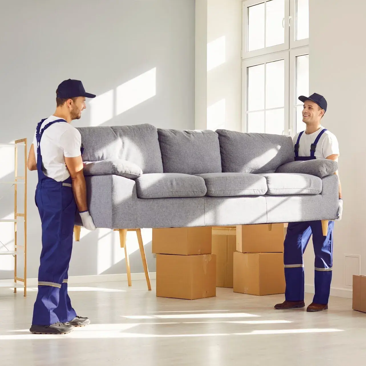 Two young delivery service workers in uniform lift heavy sofa. Men remove furniture and belongings from apartment to load in van and transport to new place. Loaders help carry couch and other things