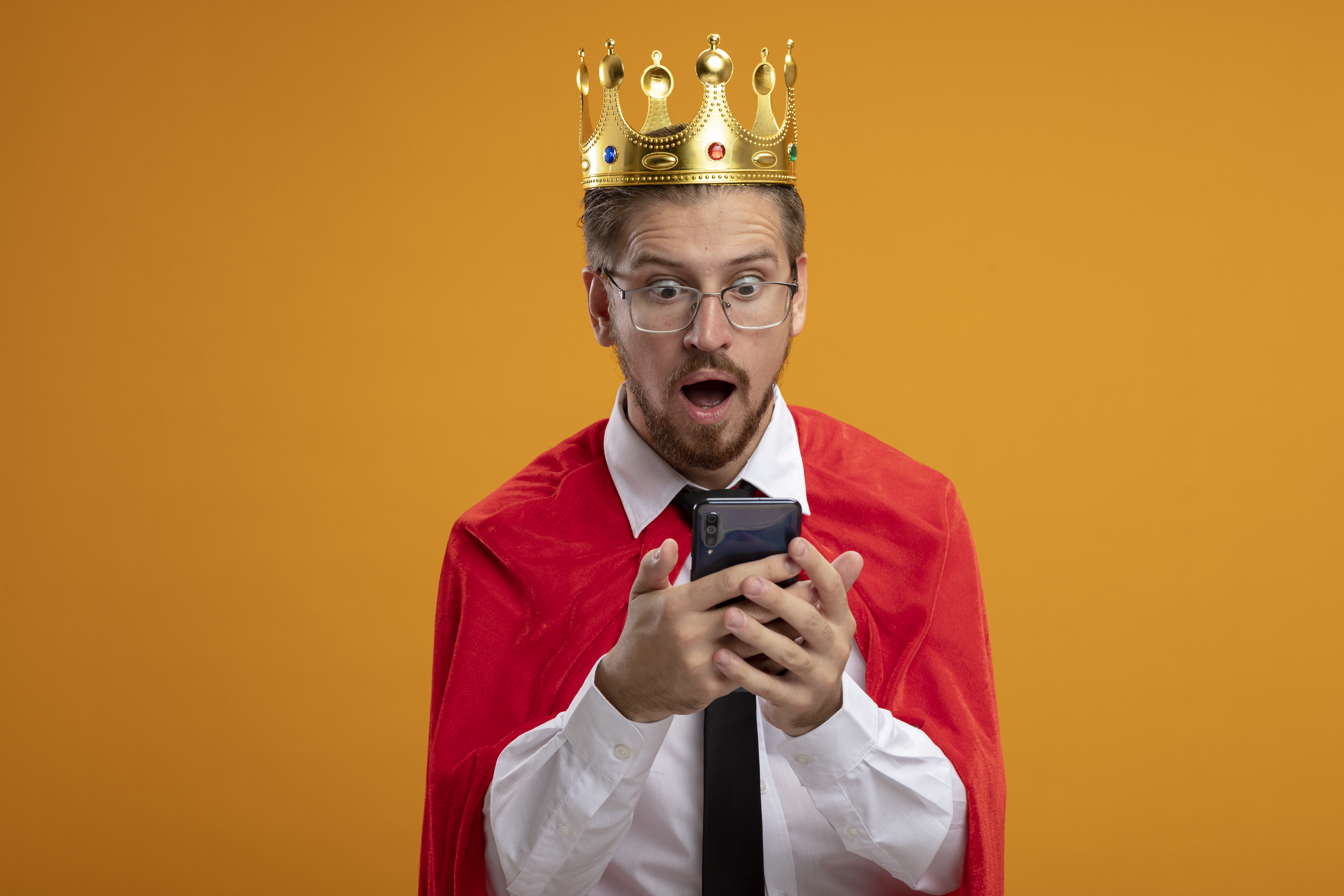 surprised-young-superhero-guy-wearing-tie-crown-with-glasses-holding-looking-phone-isolated-orange-background