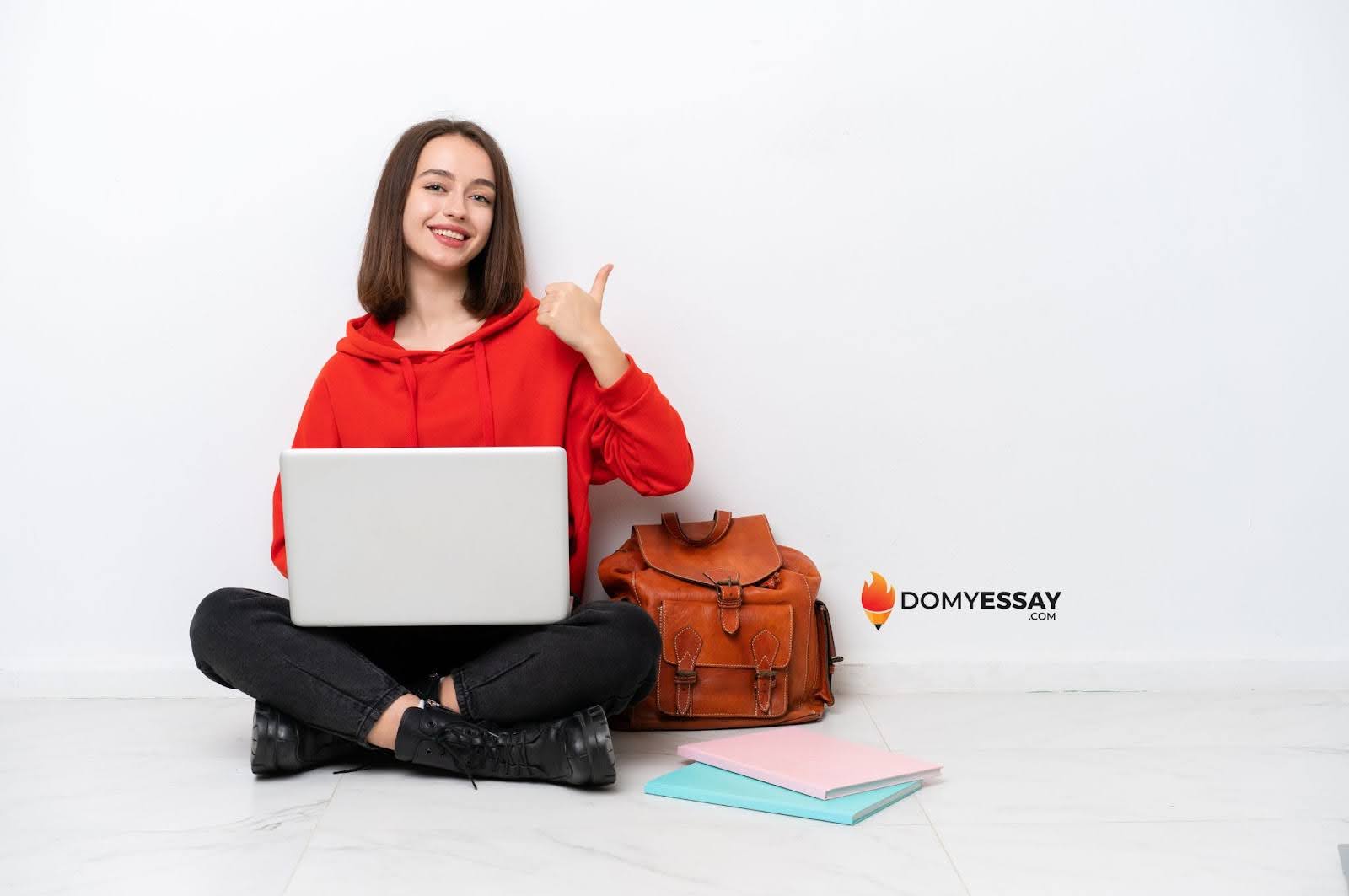 Person in a red hoodie sits on the floor with a laptop, giving a thumbs-up, next to a backpack and two closed notebooks.