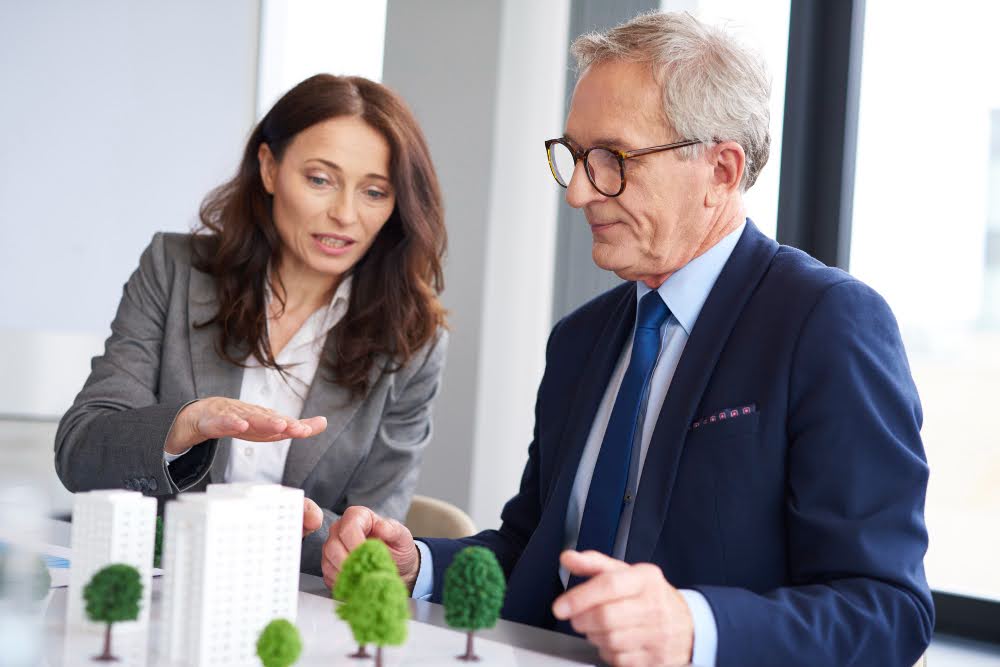 A professional woman and an older man in a business meeting, discussing a miniature urban development model.
