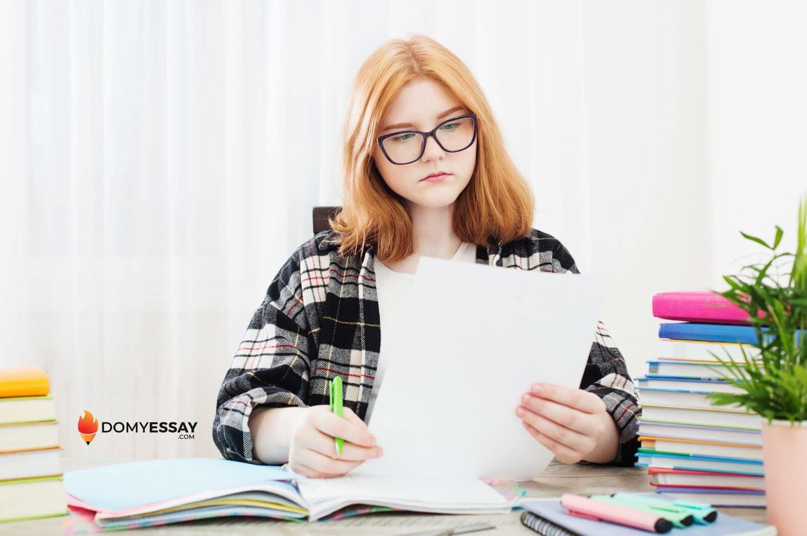 Person holds a paper and green pen at a desk, surrounded by books. Logo in the corner reads "DOMYESSAY.COM" with a flame icon.