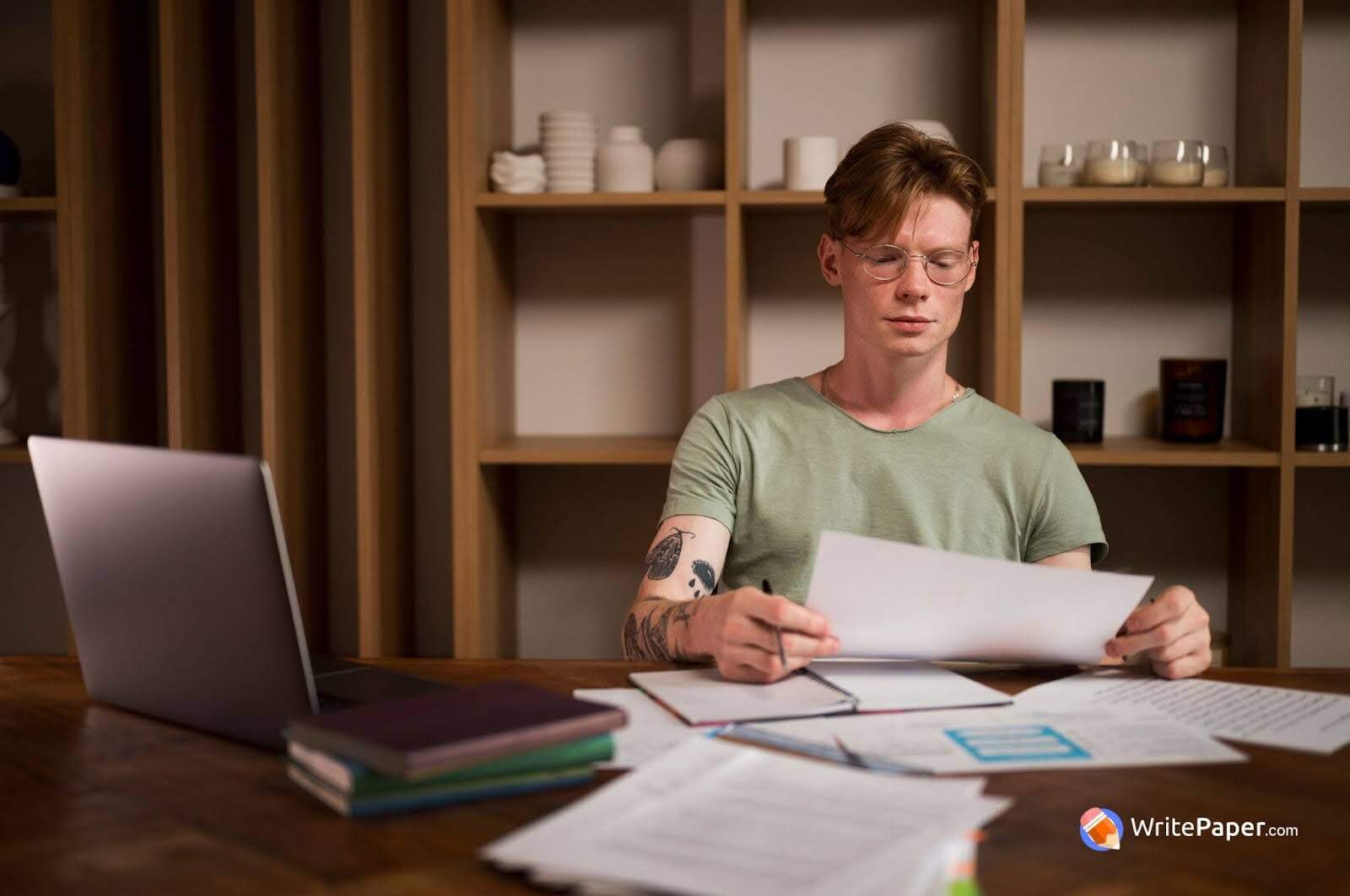 Person working at a wooden table with a laptop, books, and papers; background includes a shelf with candles and containers. Watermark reads "WritePaper.com".