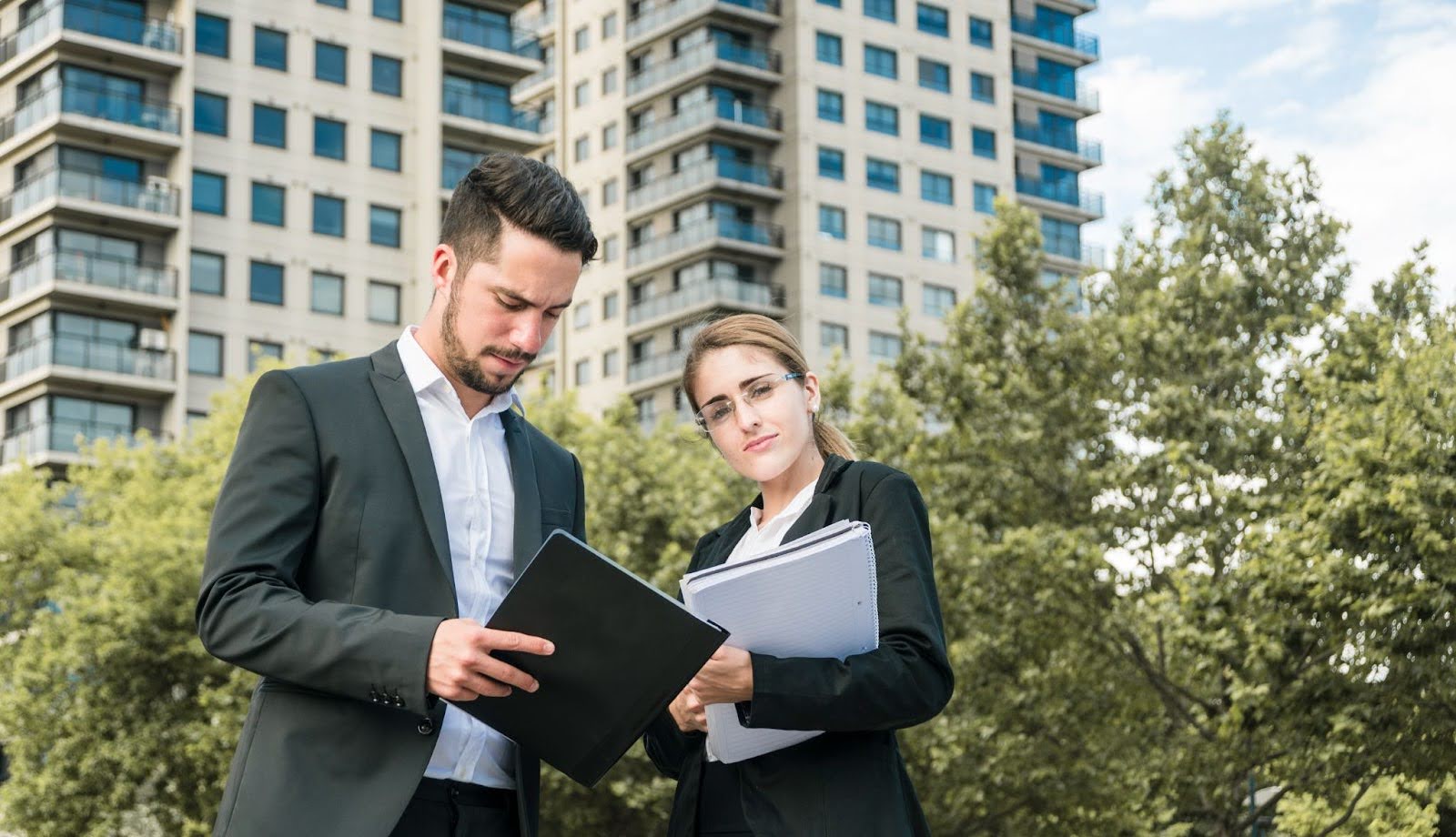 Two real estate professionals reviewing documents outdoors, with modern apartment buildings in the background.